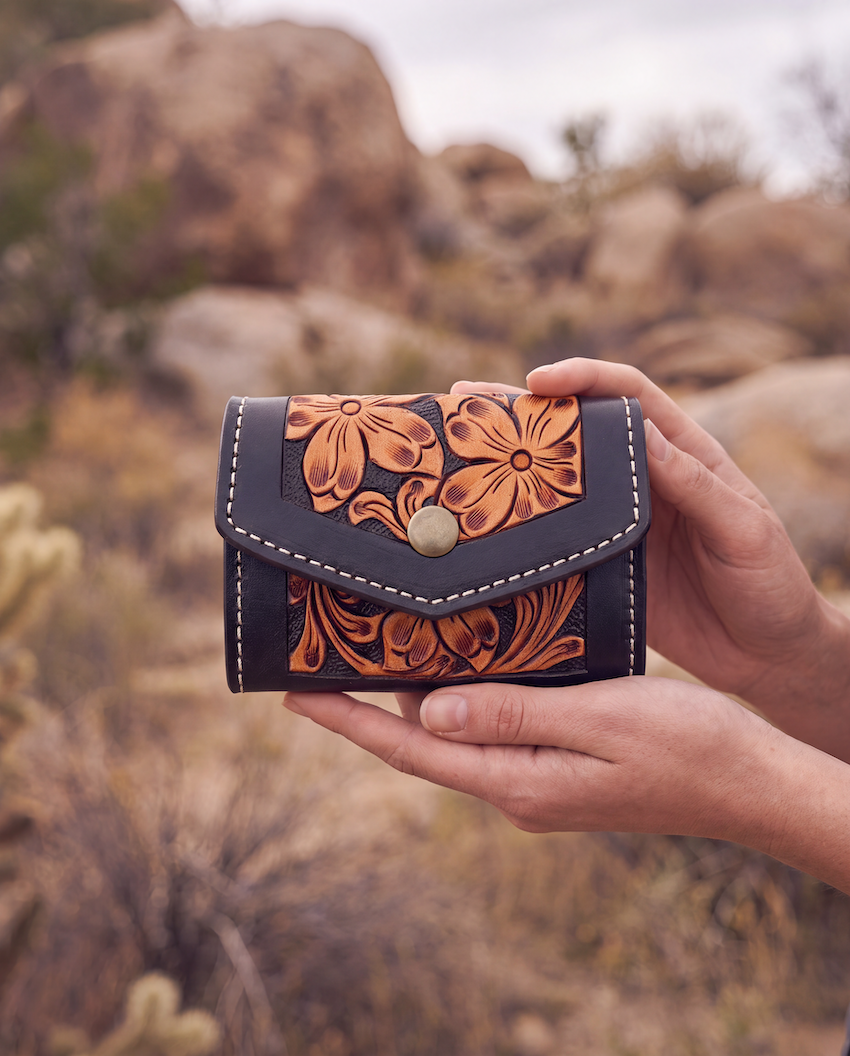 A hand holds a small tooled leather wallet in a desert landscape with rocks and cacti in the background. The wallet is dark brown with orange-brown floral tooling and white stitching.