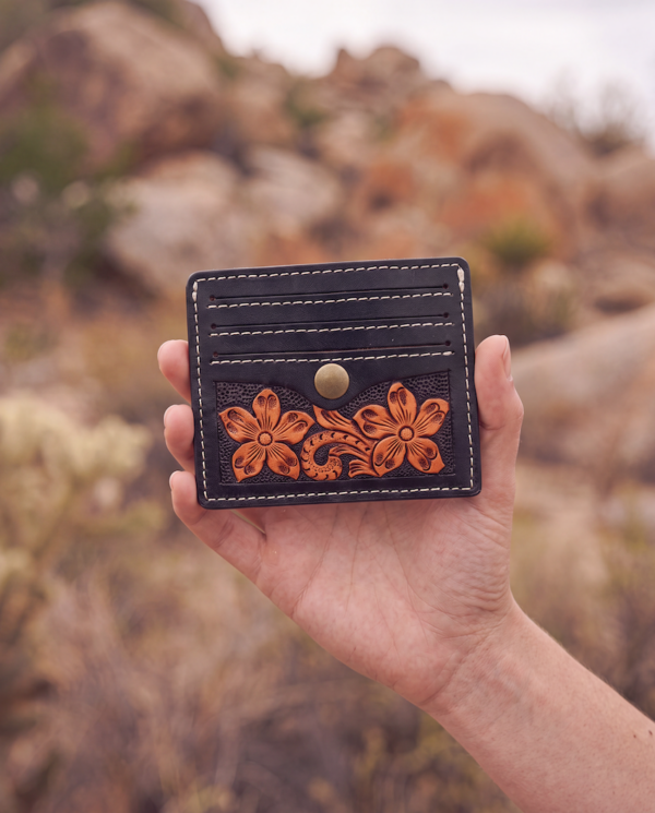 A hand holding a black leather card holder wallet with white contrast stitching and a tan floral tooled design on the front, set against a blurred outdoor desert landscape.
