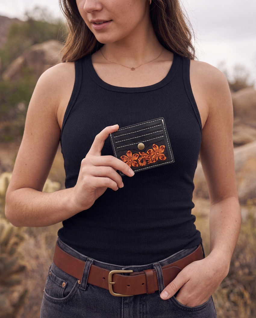 A woman wearing a black ribbed tank top and blue jeans stands outdoors in a rocky, dry landscape, holding up a small black card holder with orange floral embroidery and a gold snap button.