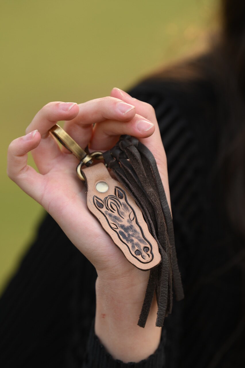 The Design Edge Tooled Leather Horse Keyring. Hand holding a leather keychain with tassels against a blurred background