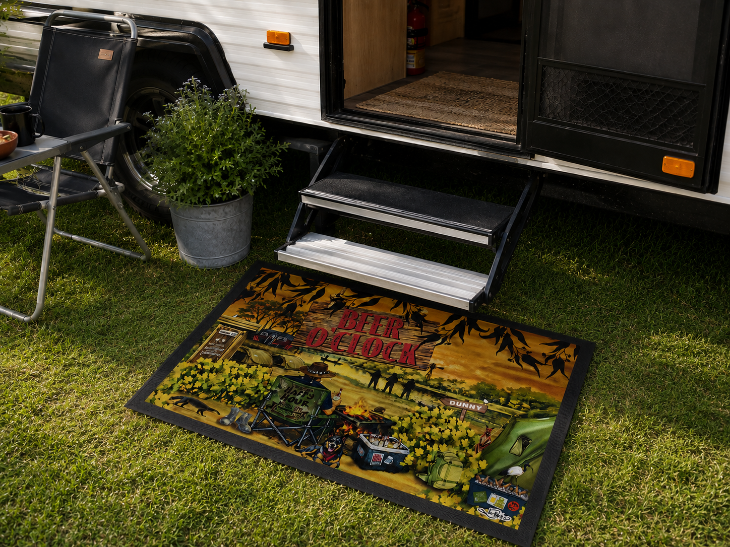 A rectangular outdoor mat with a rustic campsite illustration and the words 'Rustic Outflock' lies on green grass in front of an RV with steps extended. A folding chair and potted plant are visible nearby.