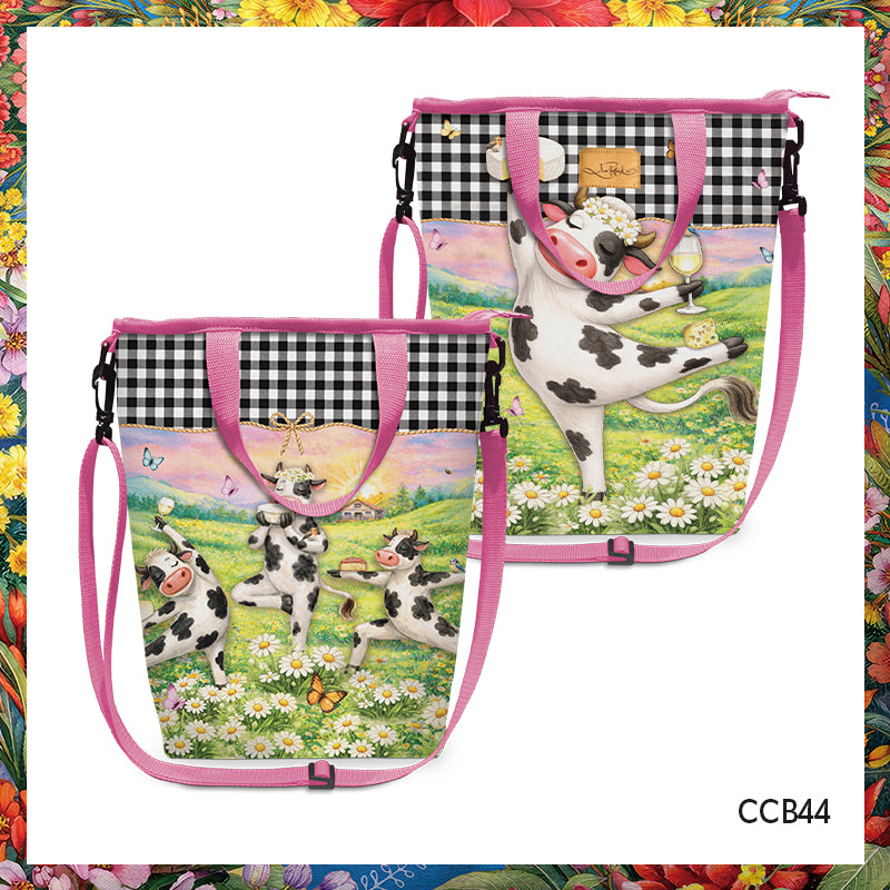 Two tote bags with pink handles and straps, featuring a black-and-white cow illustration on a white background with a floral border around the image. One bag shows two cows in a flower field, and the other shows a cow standing on a plaid section above the main illustration.