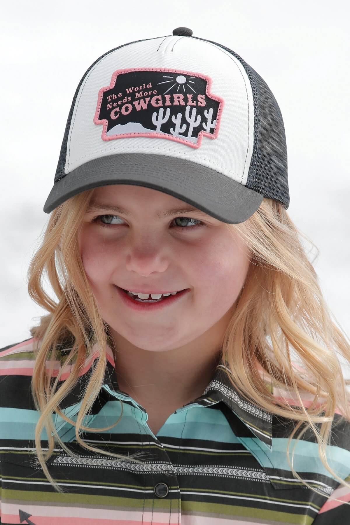 Cruel Denim Girl's The World Needs More Cowgirls Cap. A young blonde girl wearing a black mesh back trucker cap with a pink 'The World Needs More Cowgirls' woven patch on the front.
