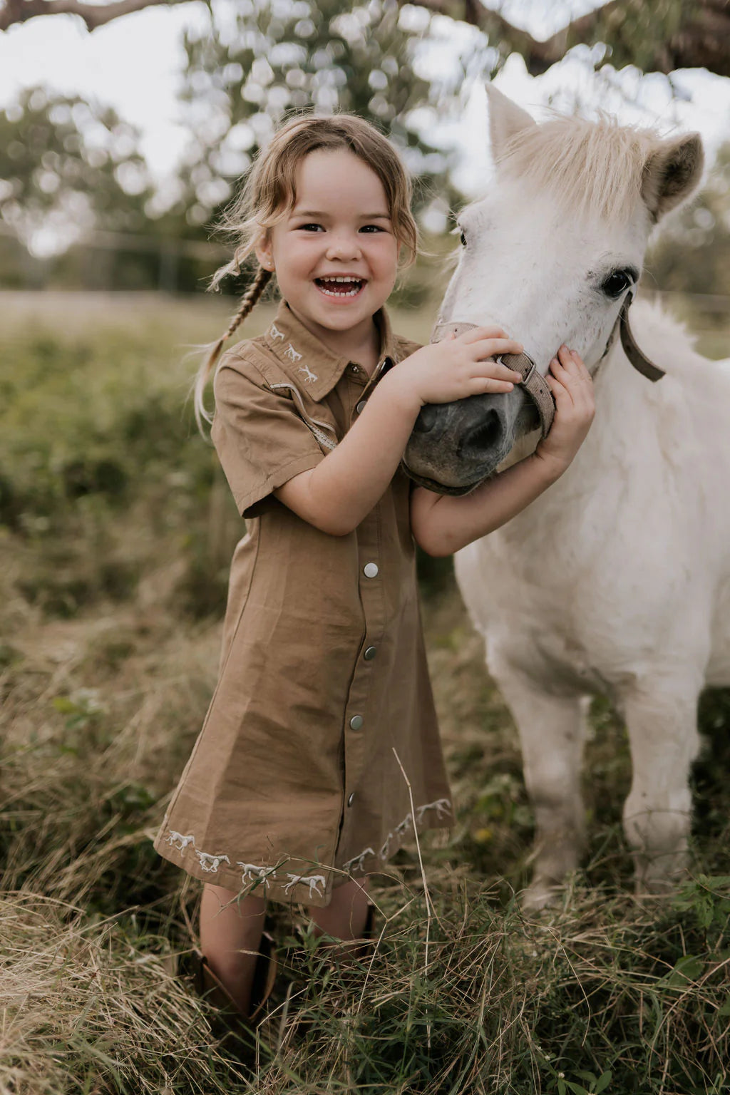Little Windmill Cheyenne Western Mocha Dress. Young girl in a brown dress hugging a small white horse in a field.