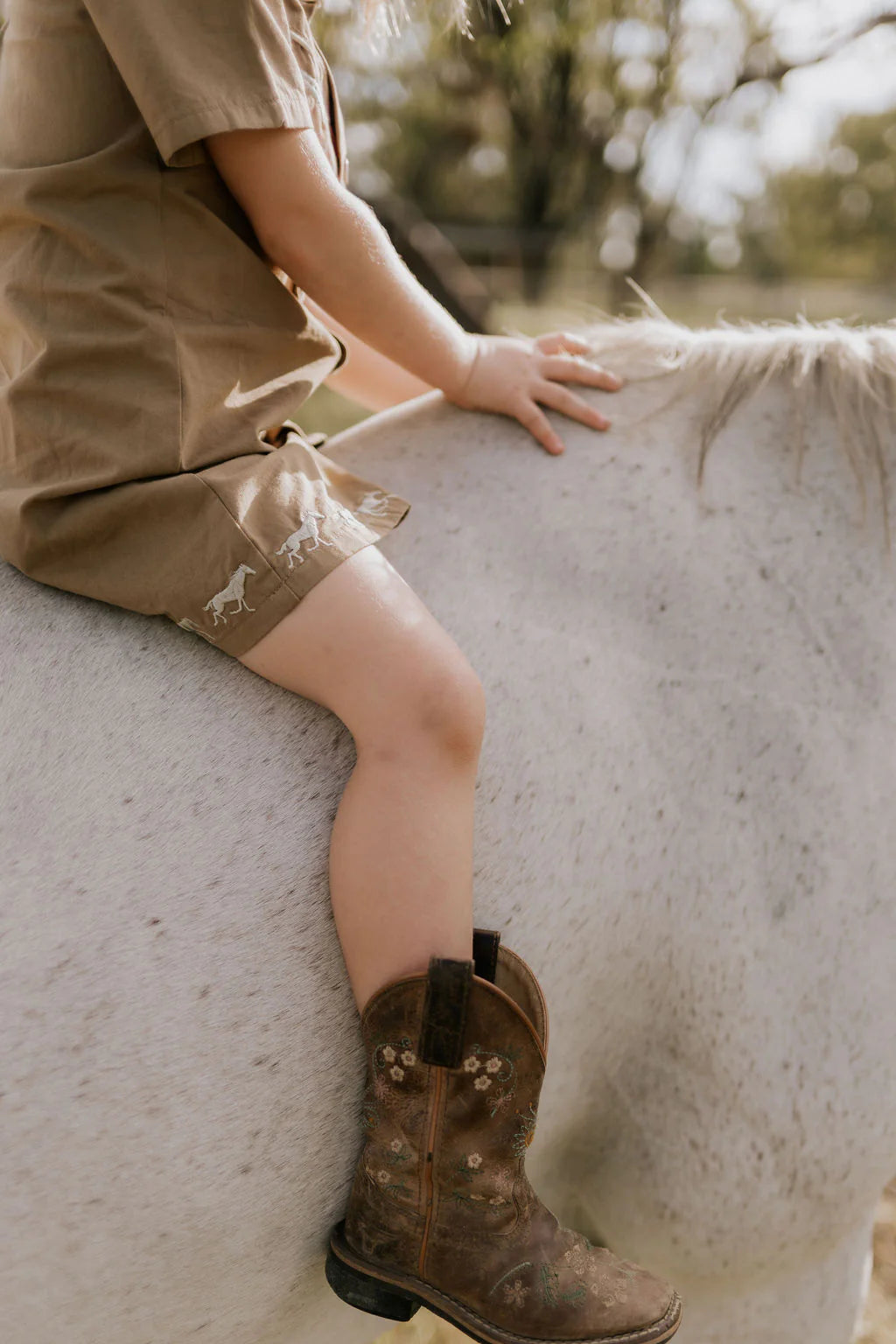 Little Windmill Cheyenne Western Mocha Dress. Person wearing brown boots and shorts sitting on a horse, with a blurred natural background.