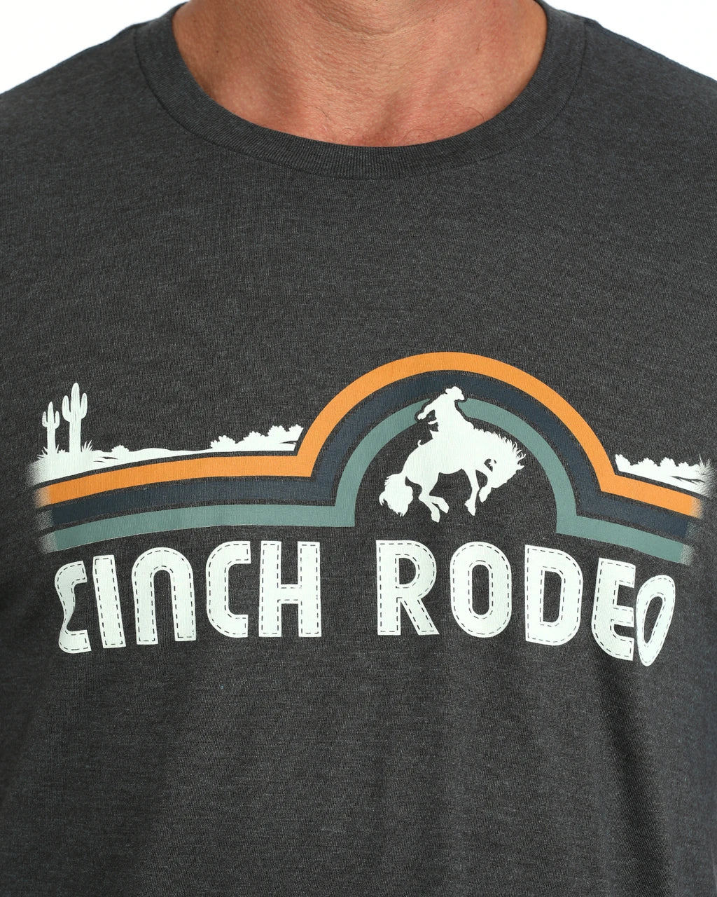 Close-up of a dark heather gray t-shirt featuring a 'Cinch Rodeo' graphic with a white cowboy on a bucking horse, an orange and teal arch, and white desert silhouettes.