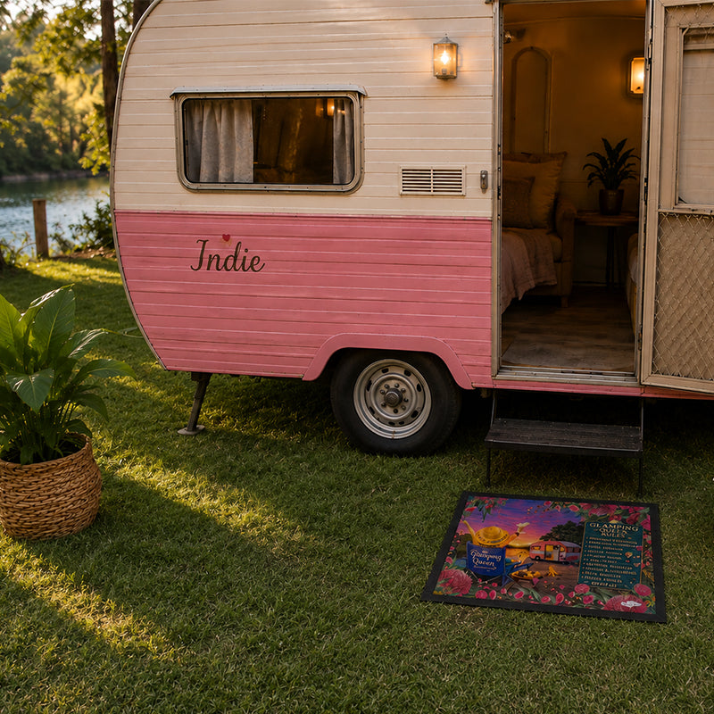 A retro-style pink and cream trailer parked on a grassy lawn beside a lake, with its door open to reveal a cozy interior. A colorful doormat sits on the grass in front, and the word 'Indie' is printed on the side of the trailer.