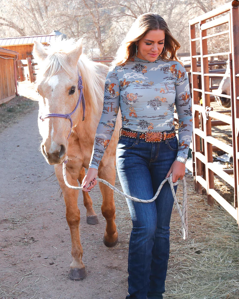 Woman wearing a patterned long-sleeve shirt with horses and nature designs, standing in front of a horse.