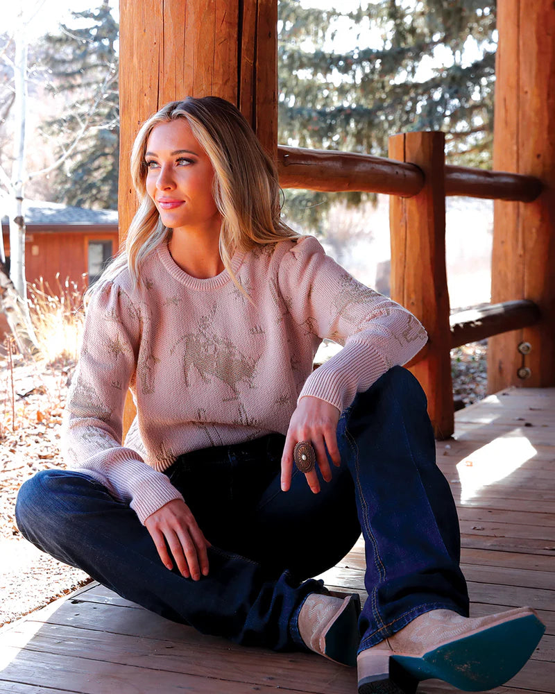 Woman sitting on a wooden deck wearing a patterned sweater and jeans.