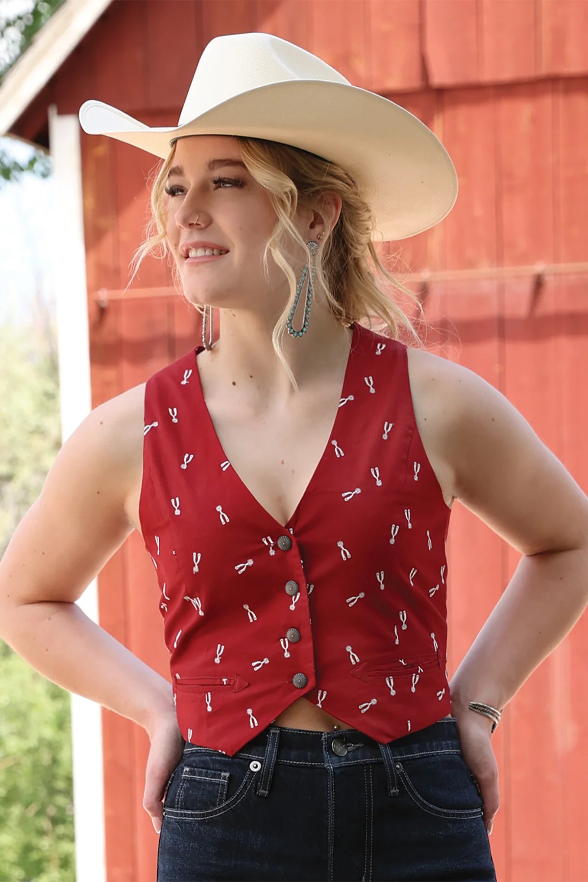 Woman wearing a red patterned vest and white cowboy hat in front of a red barn.