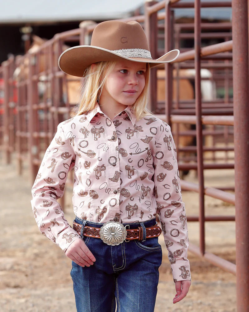 Young girl in cowboy attire with a patterned shirt and brown hat in a rodeo arena.