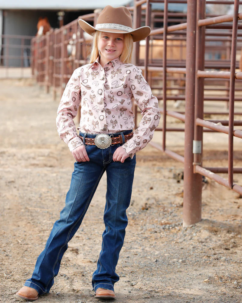 Young girl in cowboy attire with a patterned shirt and brown hat in a rodeo arena.