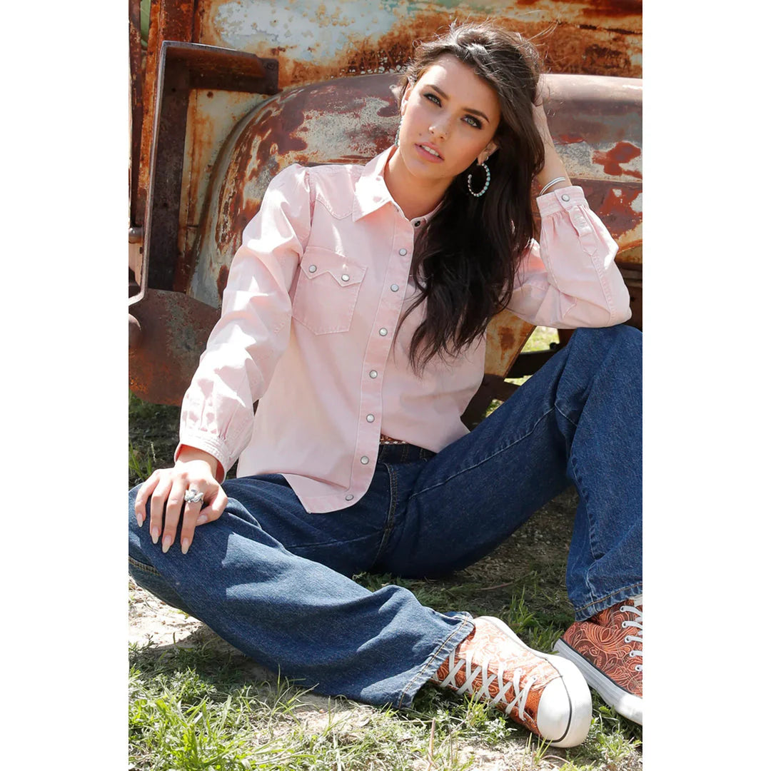 Cruel Denim Women's Pink Pearl Snap Arena Shirt. Woman sitting on the ground wearing a light pink blouse and blue jeans, with a rustic background.