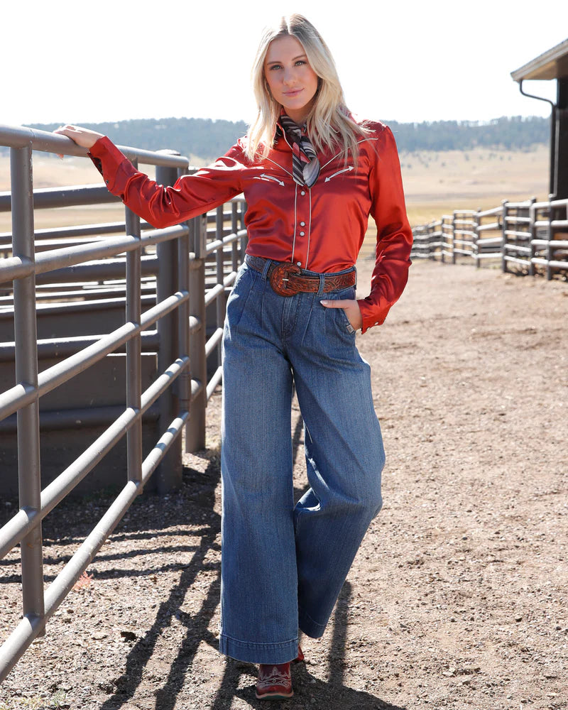 Woman in red shirt and blue jeans standing in a rural setting with a fence and open field.