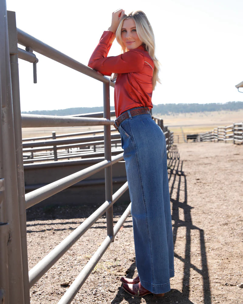 Woman in red shirt and blue jeans standing in a rural setting with a fence and open field.