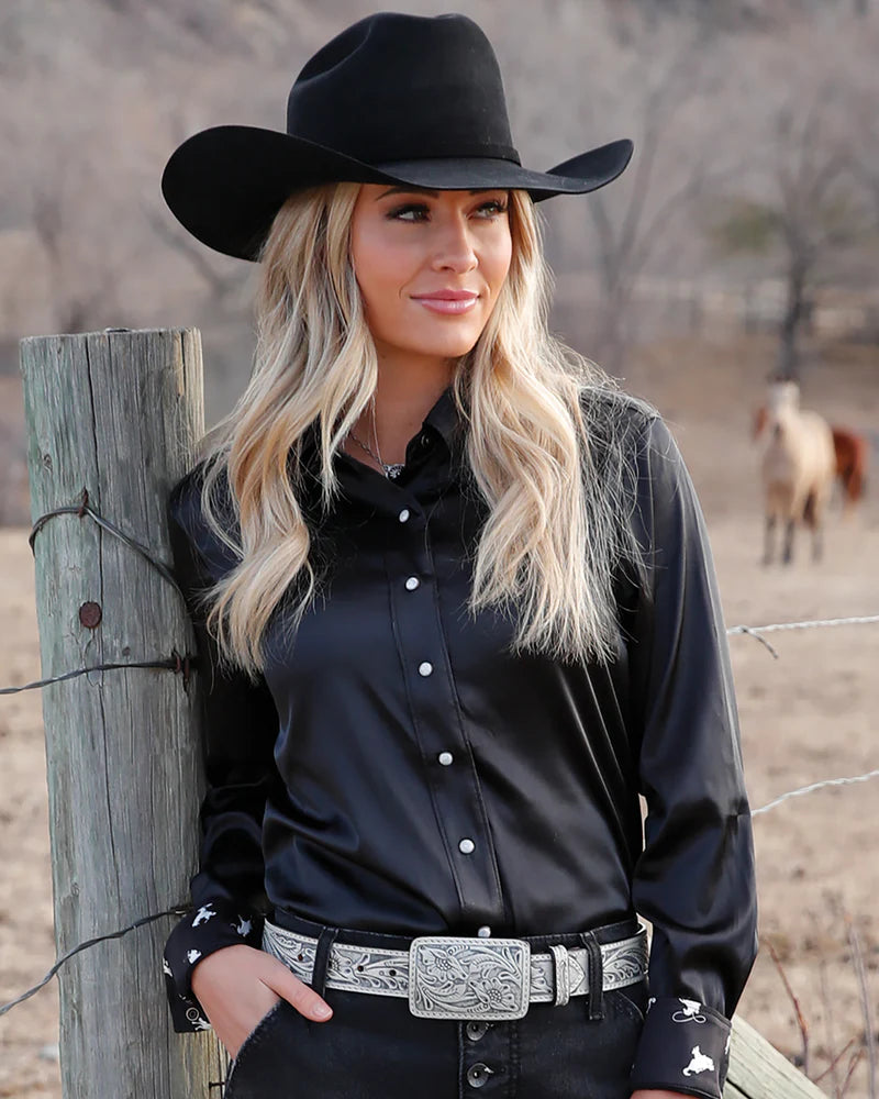Woman wearing a black cowboy hat and shirt in a rural setting with a horse in the background.