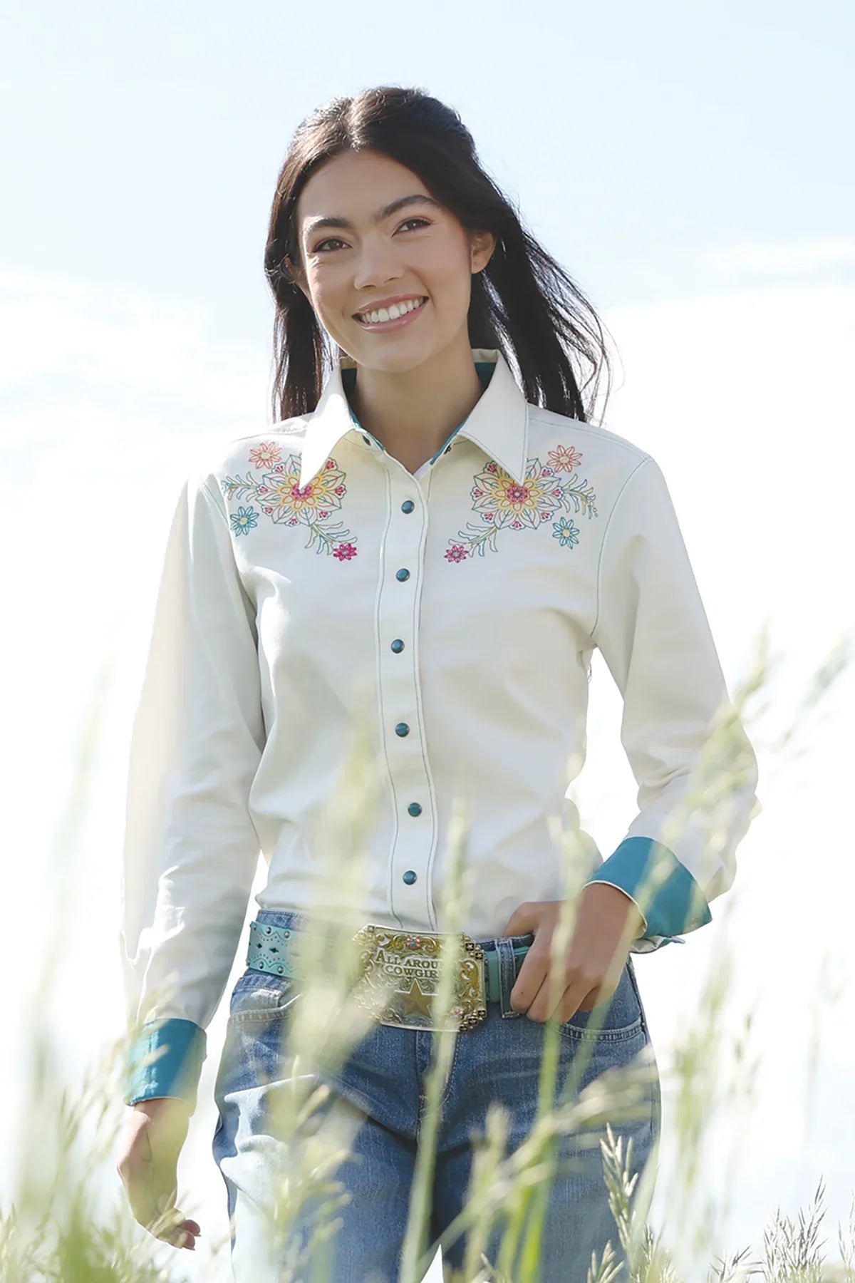 Woman wearing a white embroidered shirt and blue jeans standing in a field.