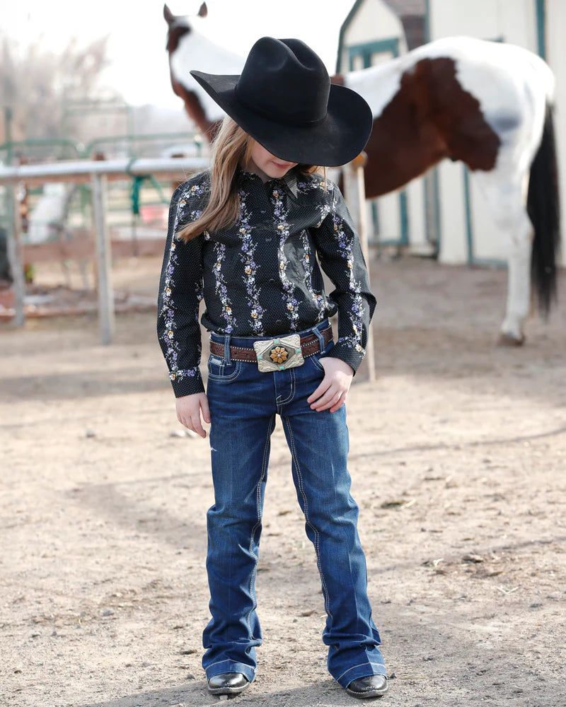 Child wearing a black cowboy hat and patterned shirt with a horse in the background