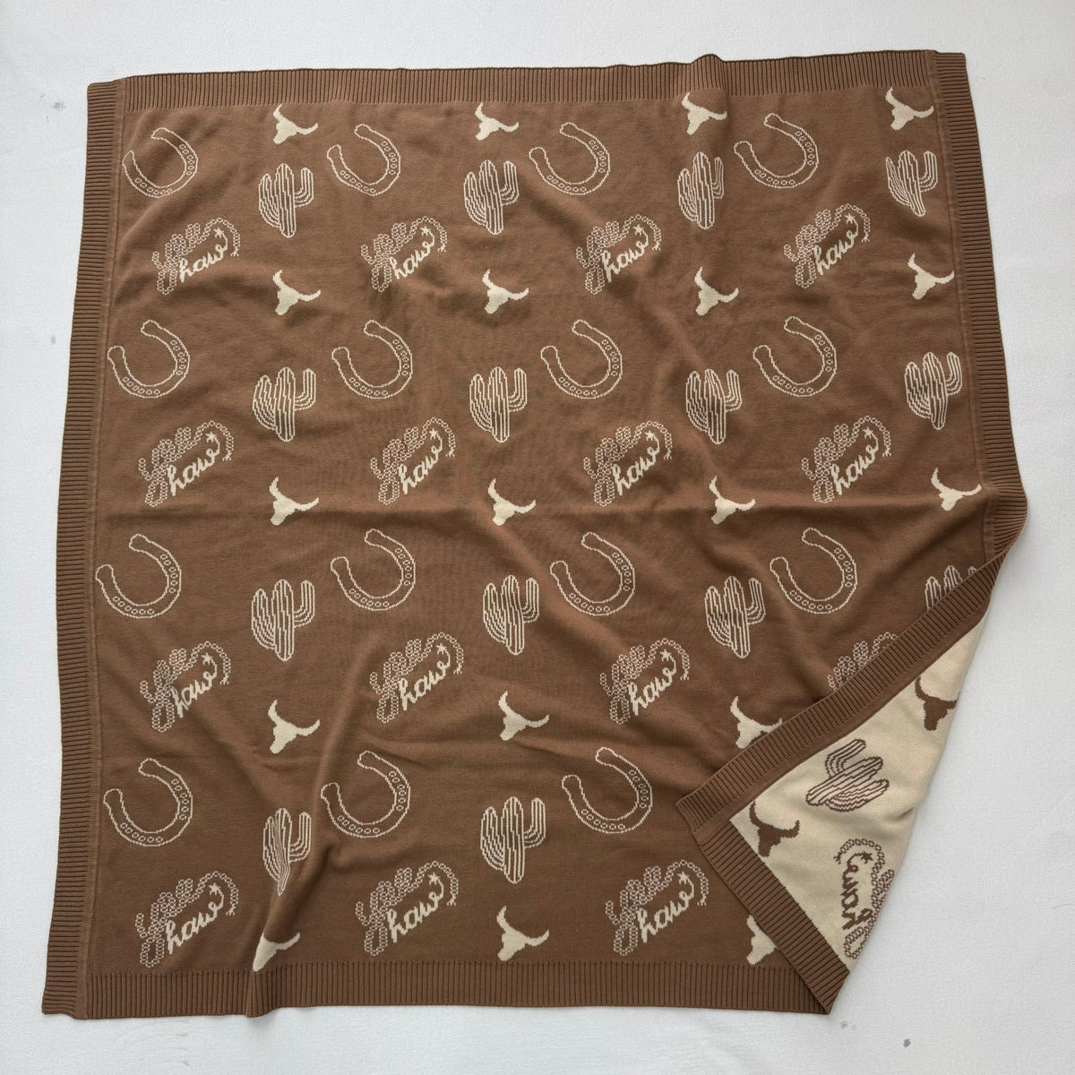 A square brown knit blanket featuring a repeating pattern of white cacti, horseshoes, longhorns, and the words 'The Cow' in script, laid flat on a white background with one corner folded over to show the reverse side.