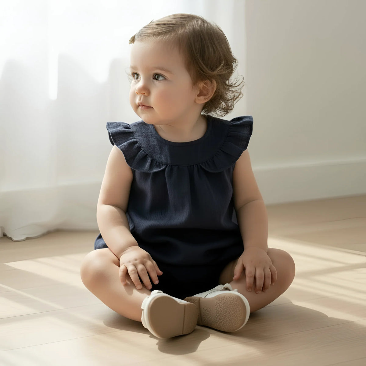 A toddler girl with light brown hair sits cross-legged on a light wood floor in a bright room, wearing a sleeveless navy blue ruffled top and beige baby shoes.