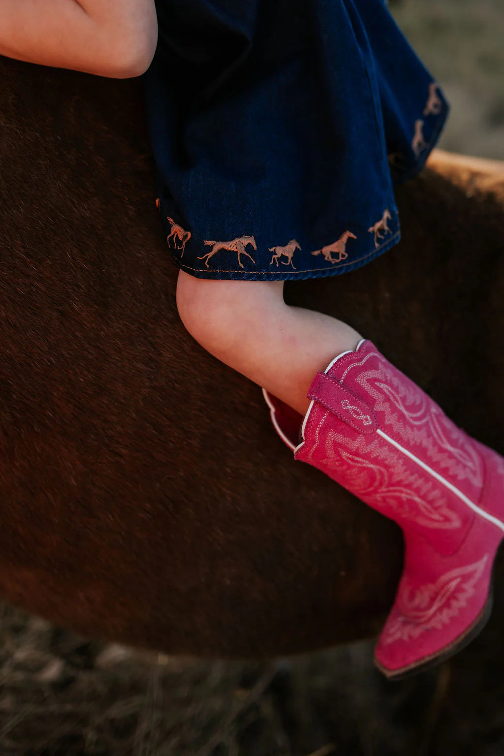 Little Windmill Girl's Dekota Dark Horse Denim Dress. Person wearing pink cowboy boot and dark blue pants with horse patterns on a horse.
