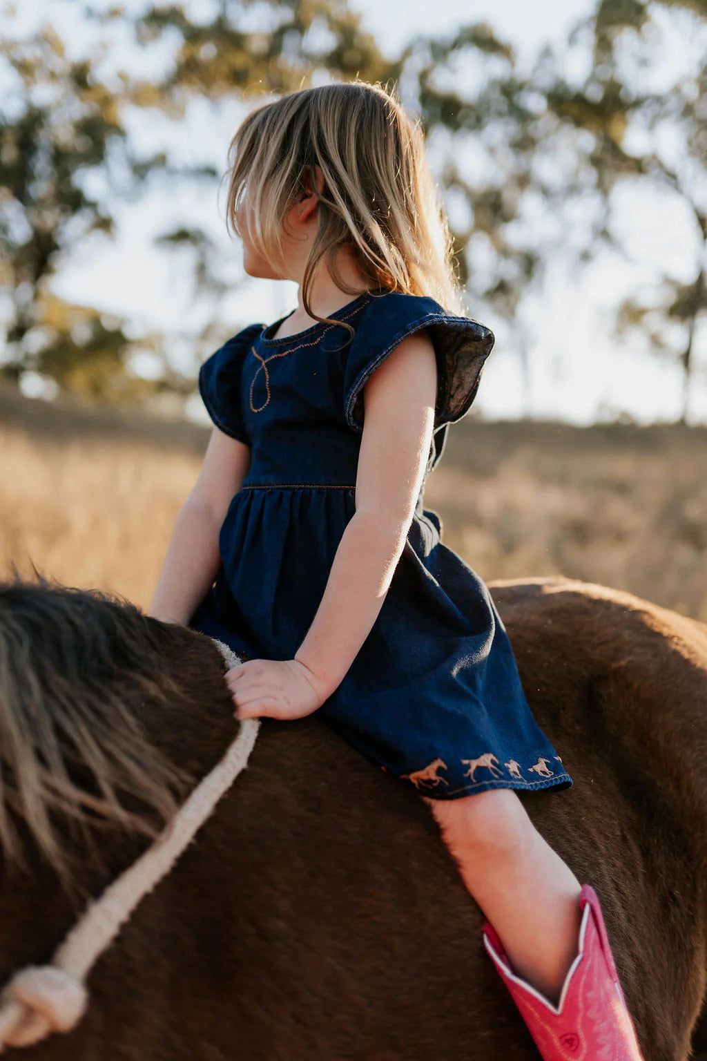 Little Windmill Girl's Dekota Dark Horse Denim Dress. Young girl in a blue dress riding a horse in an outdoor setting