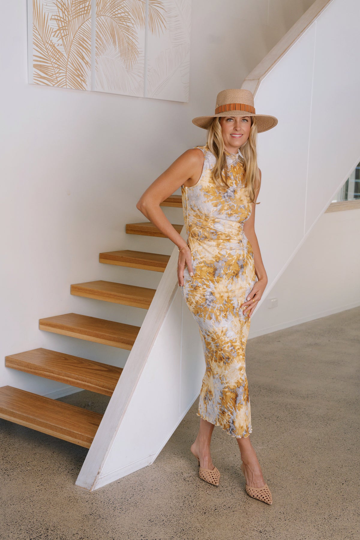 A woman wearing a sleeveless yellow and white floral midi dress, a wide-brimmed straw hat, and beige pointed flats, posing next to a wooden staircase in a modern white-walled interior.