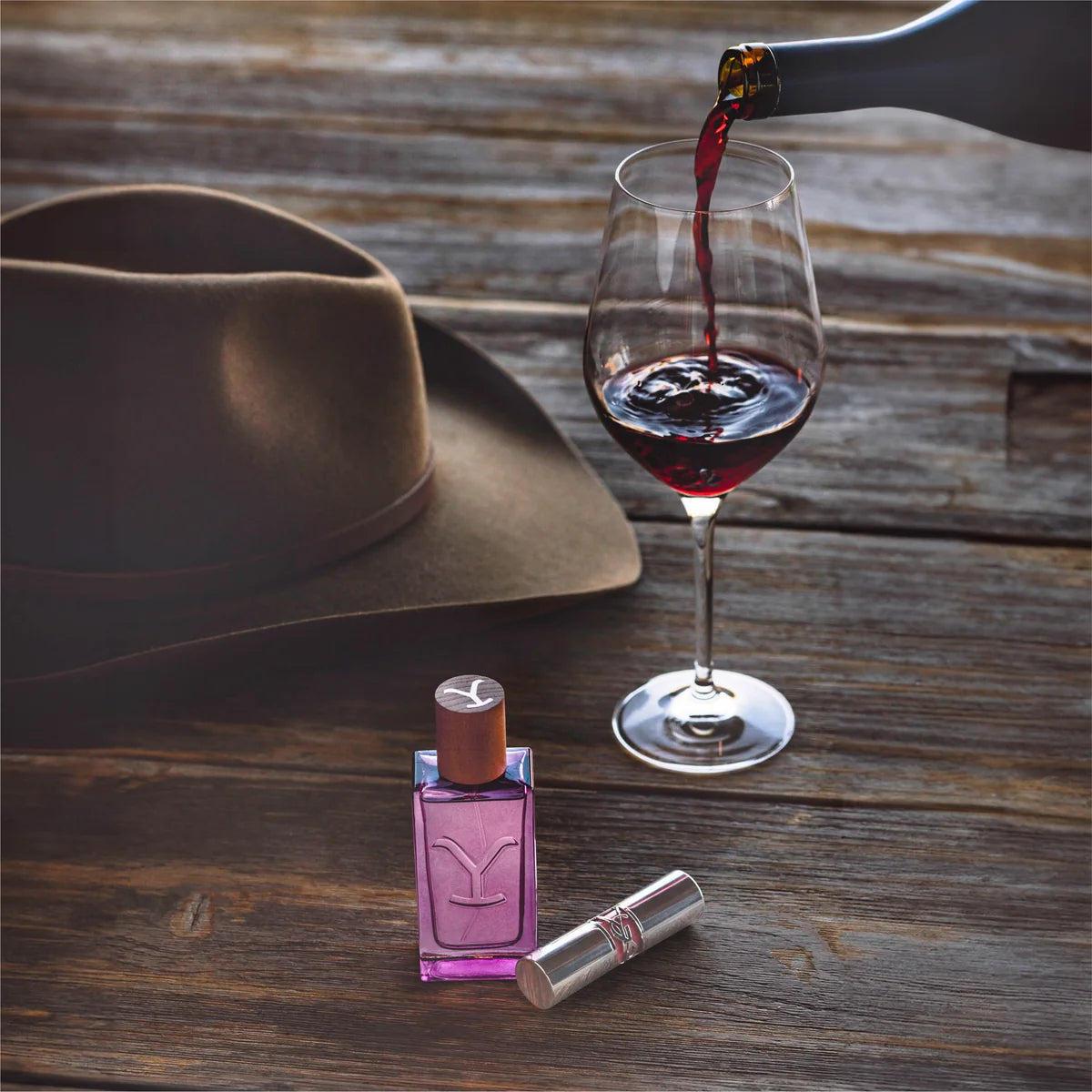 Wine being poured into a glass with a hat and perfume bottle on a wooden surface