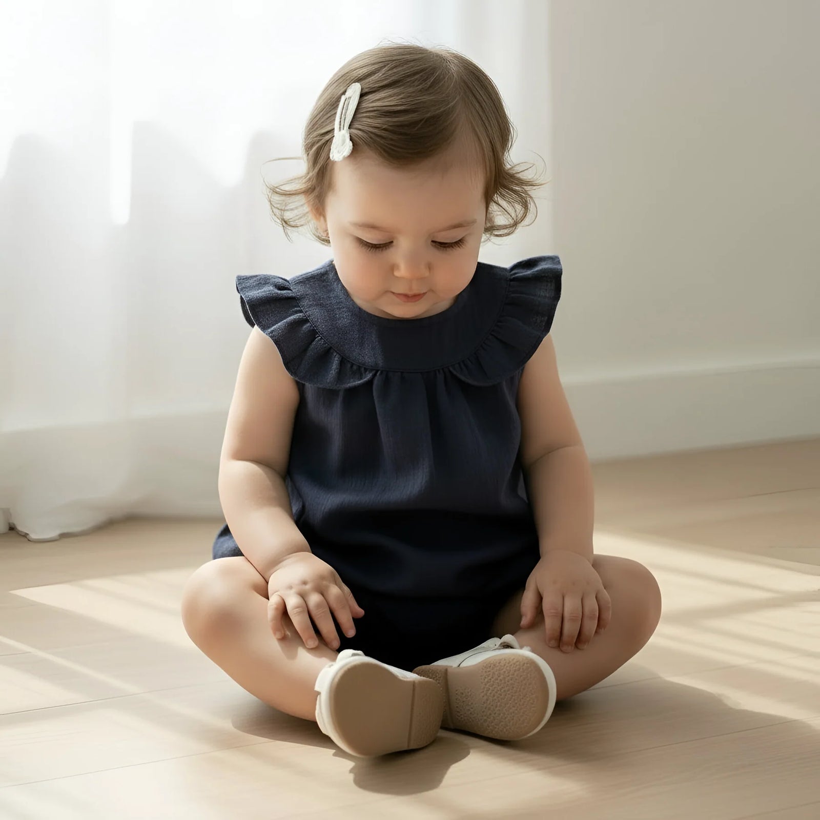 A toddler girl sitting cross-legged on a light wooden floor, wearing a sleeveless navy blue ruffled top and beige baby shoes, with a white hair clip in her hair.