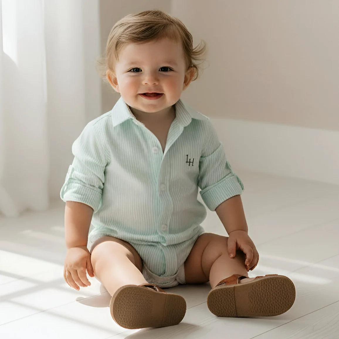 Baby wearing a light green striped shirt and brown shoes sitting on a white floor.