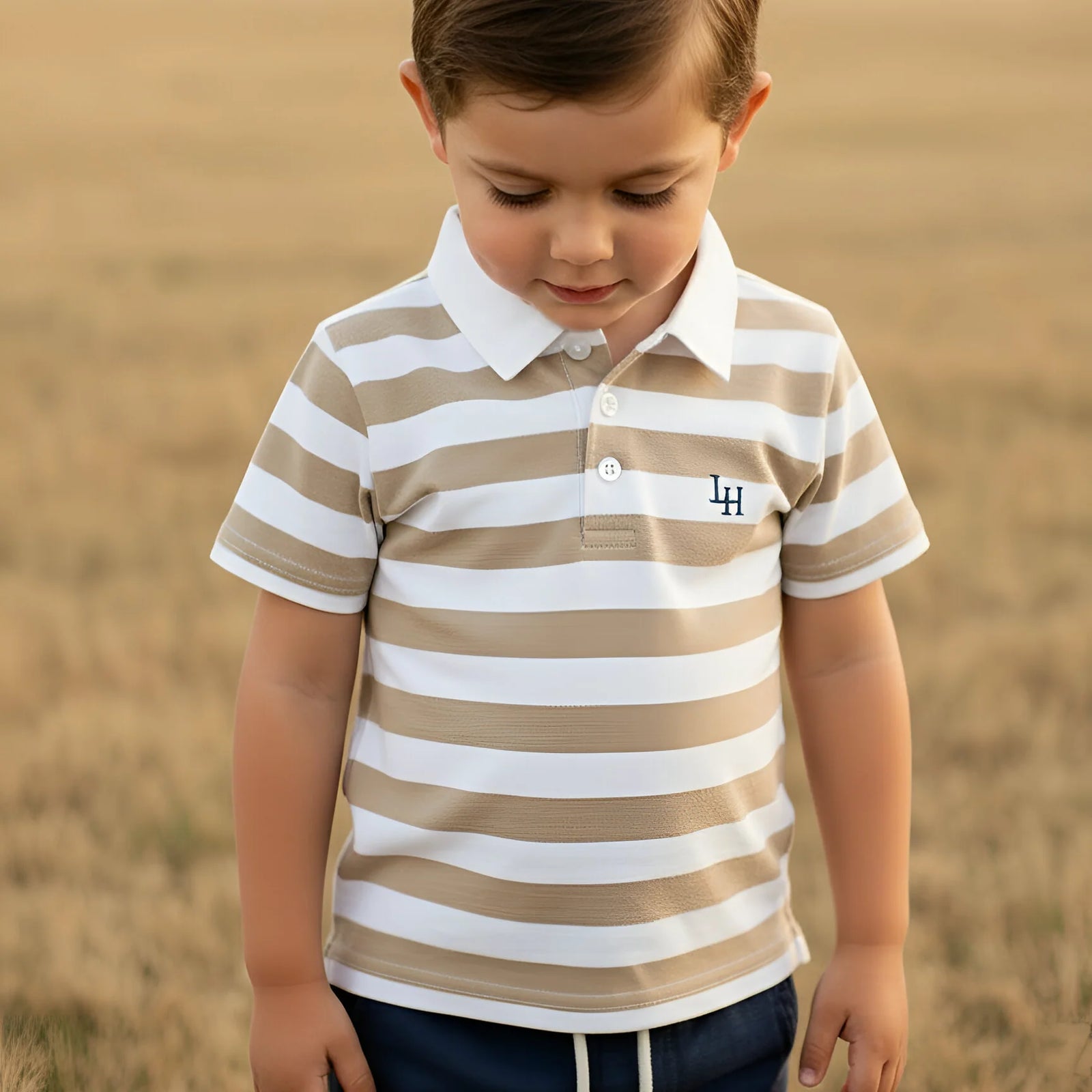 A young boy wearing a beige and white horizontally striped polo shirt with a white collar and a small embroidered 'LA' logo on the chest, standing in a dry, golden field.