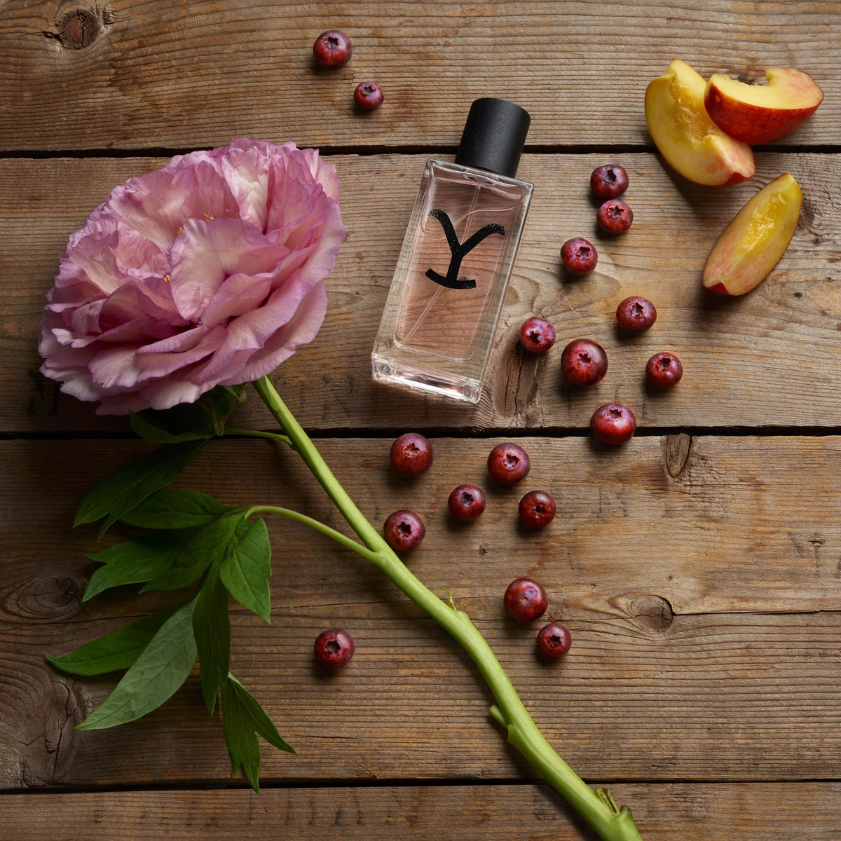 A clear rectangular perfume bottle with a black cap and a stylized black 'Y' logo on the front, arranged on a rustic wooden surface with a pink peony flower, green leaves, scattered cranberries, and sliced peach.