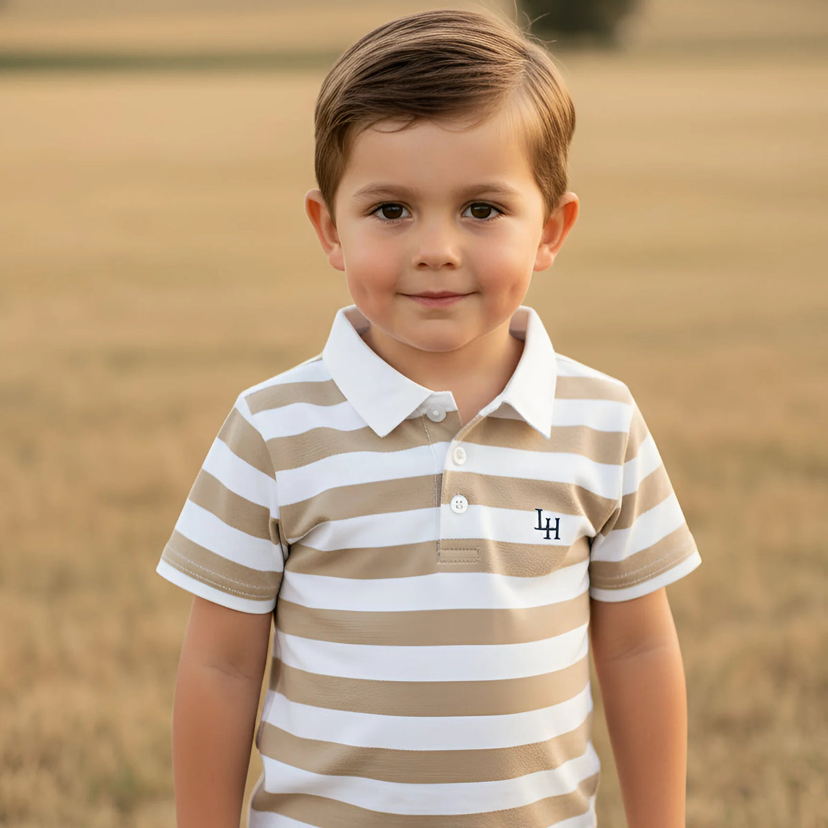 A young boy wearing a beige and white horizontally striped polo shirt with a white collar and a small embroidered 'LH' logo on the chest, standing in a blurred golden field.