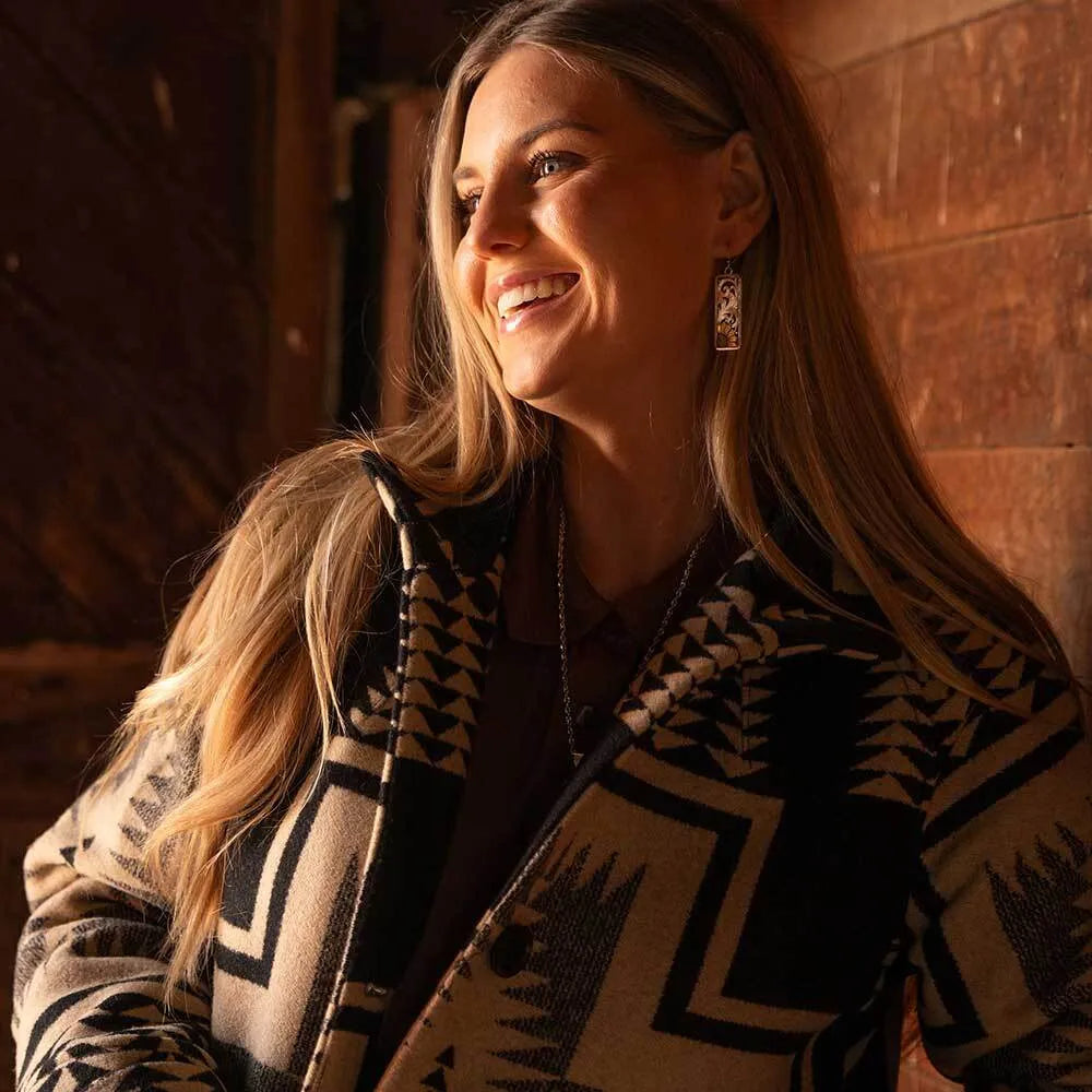 A woman smiling and looking to the side, wearing a black and beige patterned knit cardigan in warm, rustic indoor lighting.