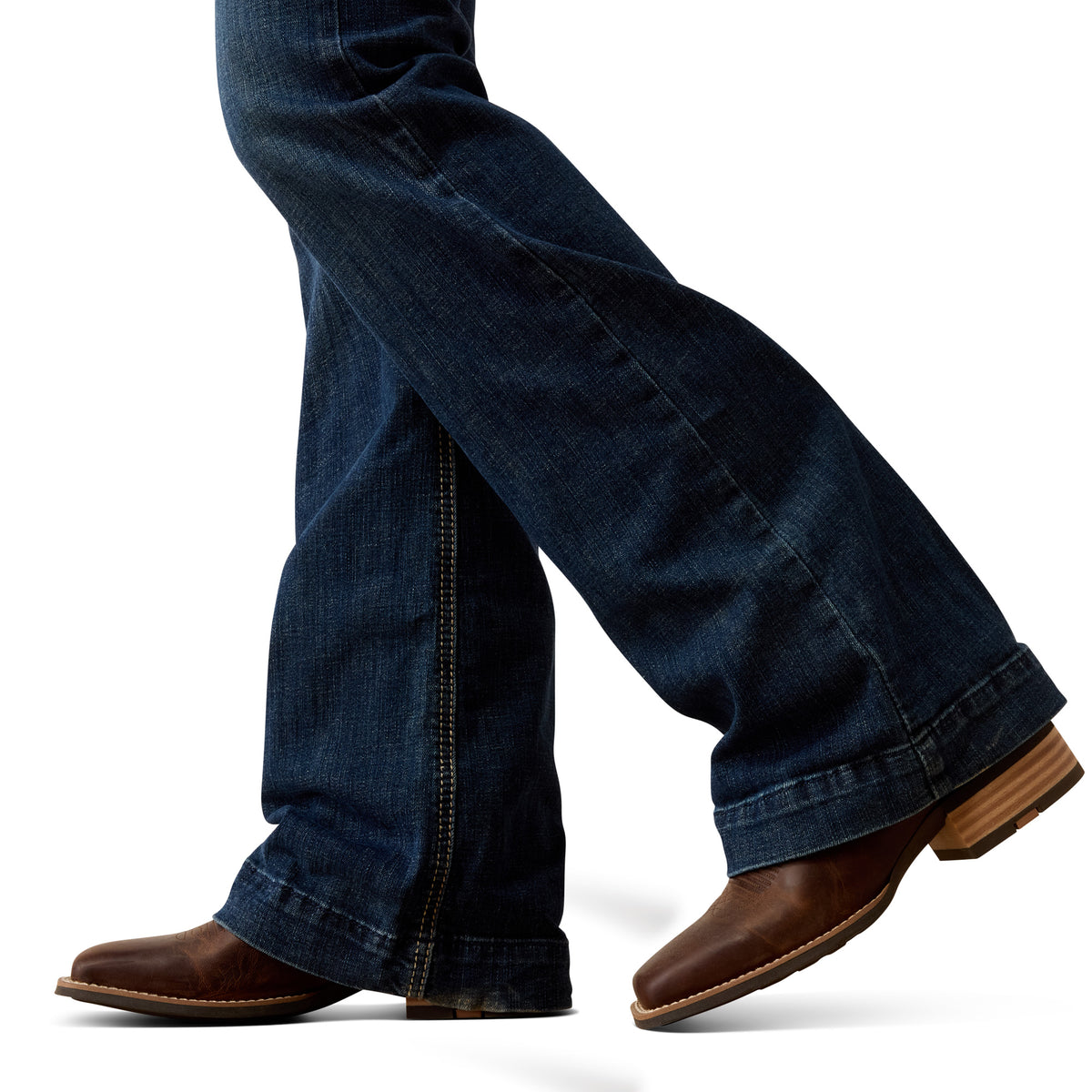 Close-up of a person's legs wearing dark blue flared jeans and brown leather boots against a white background.