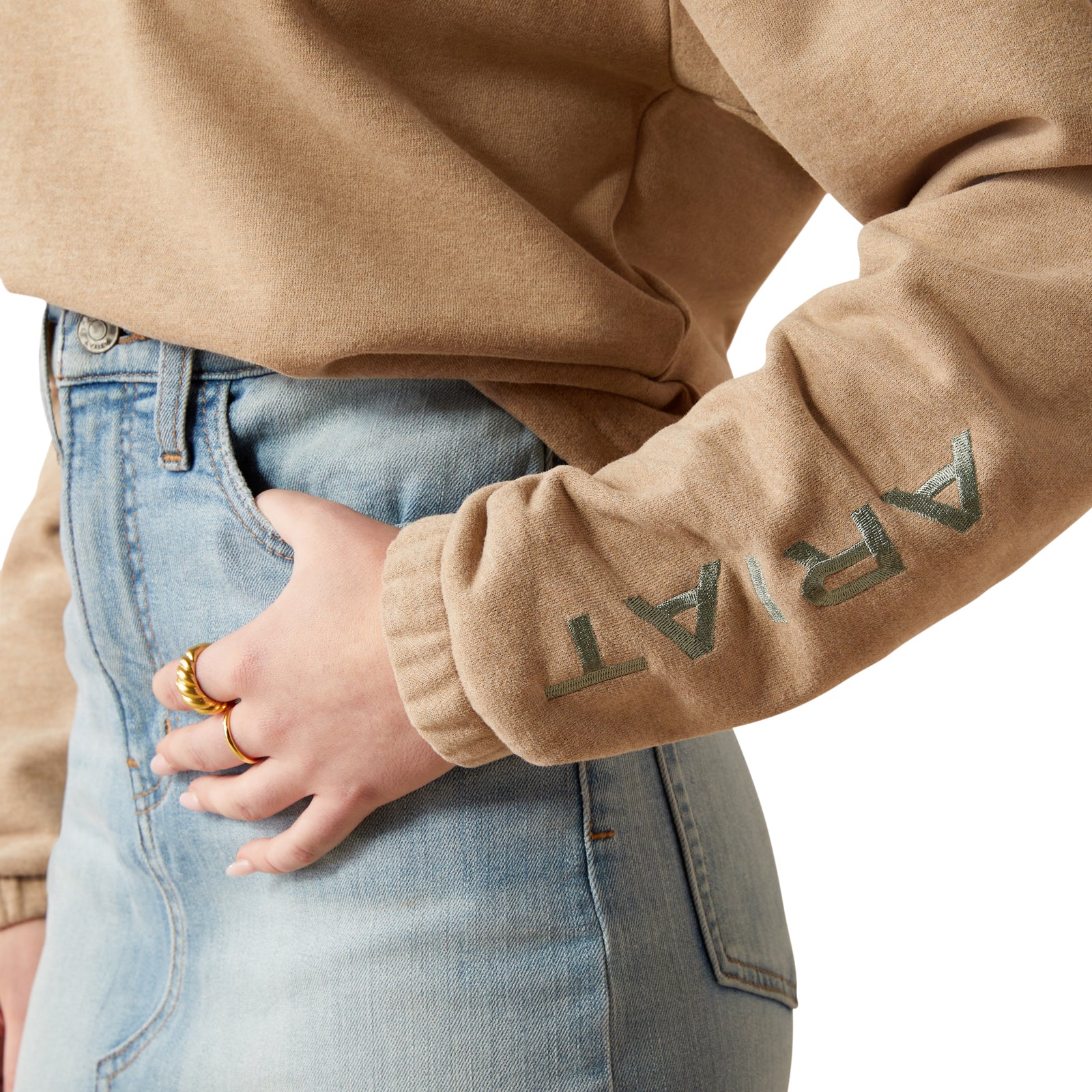 Ariat Women's Essential Logo HoodieA woman wearing a beige hoodie with the brand name 'Ariat' on the sleeve, paired with a brown cowboy hat and blue jeans. 