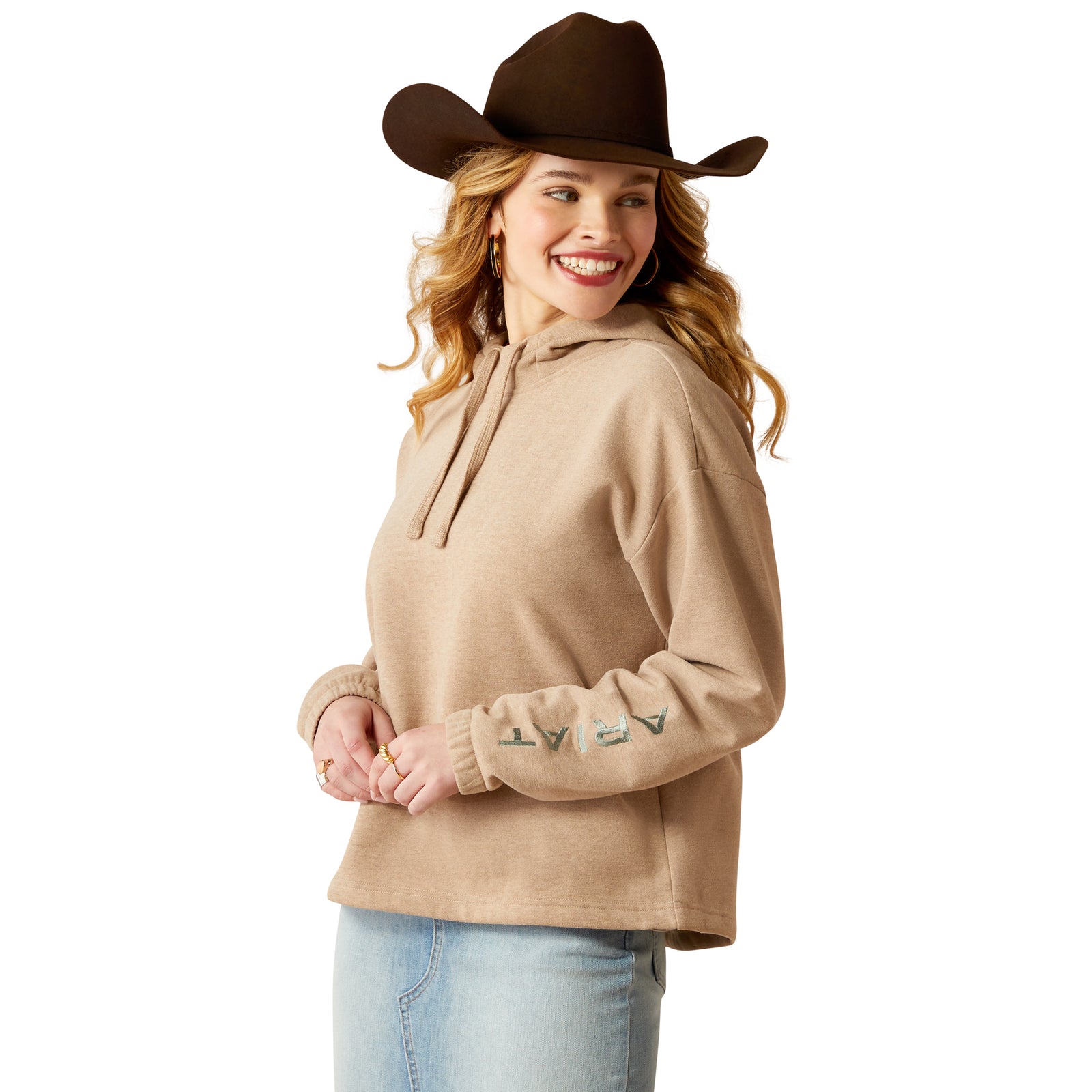 Ariat Women's Essential Logo HoodieA woman wearing a beige hoodie with the brand name 'Ariat' on the sleeve, paired with a brown cowboy hat and blue jeans. 
