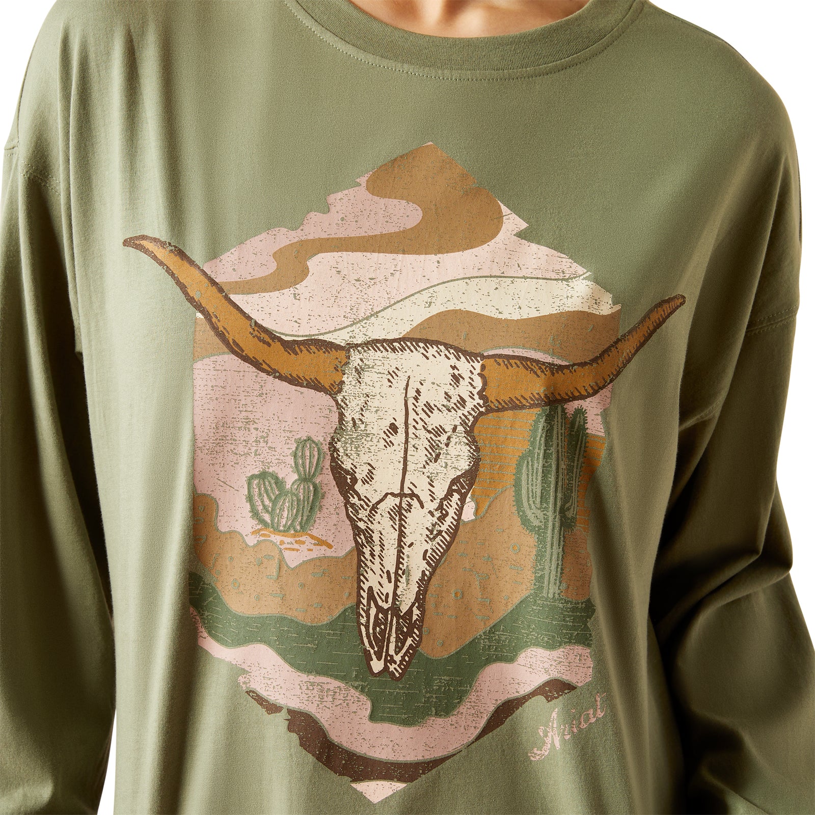 Ariat Women's Wanderer L/S T-Shirt. A woman wearing a long-sleeved green t-shirt with a graphic print of a cow's skull, paired with a brown cowboy hat and blue jeans.