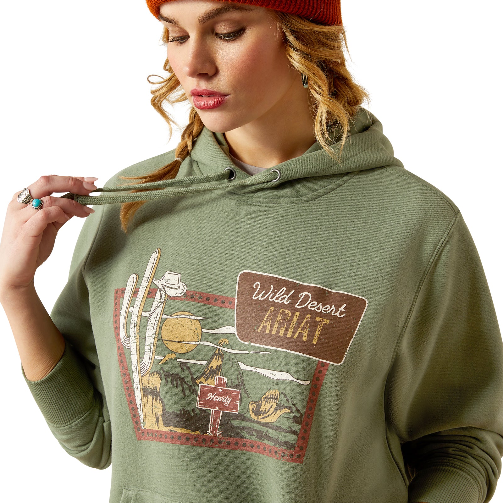 Ariat Women's Route 66 Hoodie. A woman wearing a green hoodie with a graphic print, paired with jeans and an orange beanie.