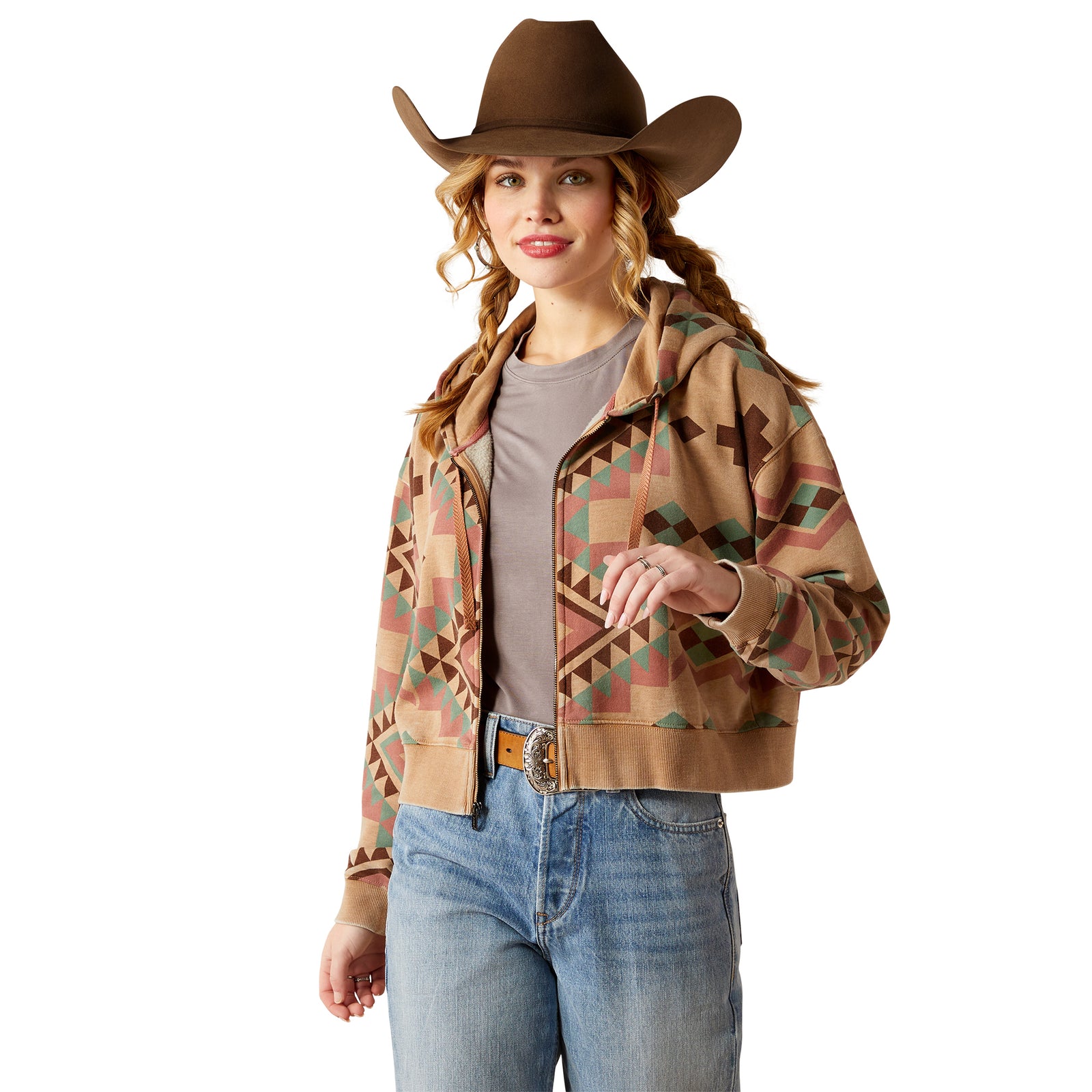 Ariat Women's Sunset Spark Crop Hoodie. A woman wearing a multicolored geometric patterned crop hoodie with a hood, paired with a gray shirt and blue jeans, accessorized with a brown hat.