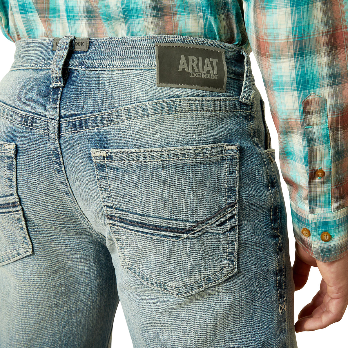 Ariat Men's M4 Thomas Relaxed Boot Cut Jean. Person wearing blue jeans with Ariat logo, plaid shirt visible