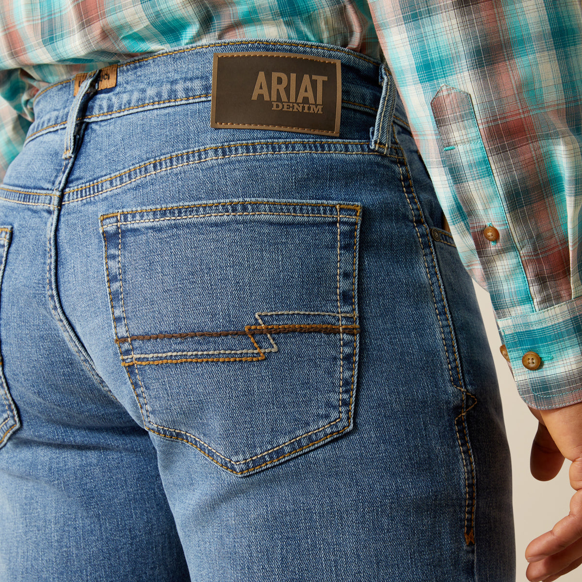 Ariat Men's M4 Pickett Relaxed Boot Cut Jean. Close-up of blue jeans with Ariat denim label and plaid shirt.