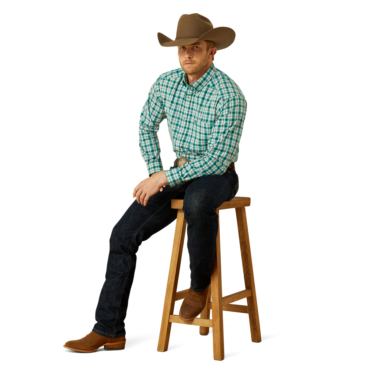 Man wearing a green plaid shirt, dark jeans, and a brown cowboy hat sitting on a wooden stool.