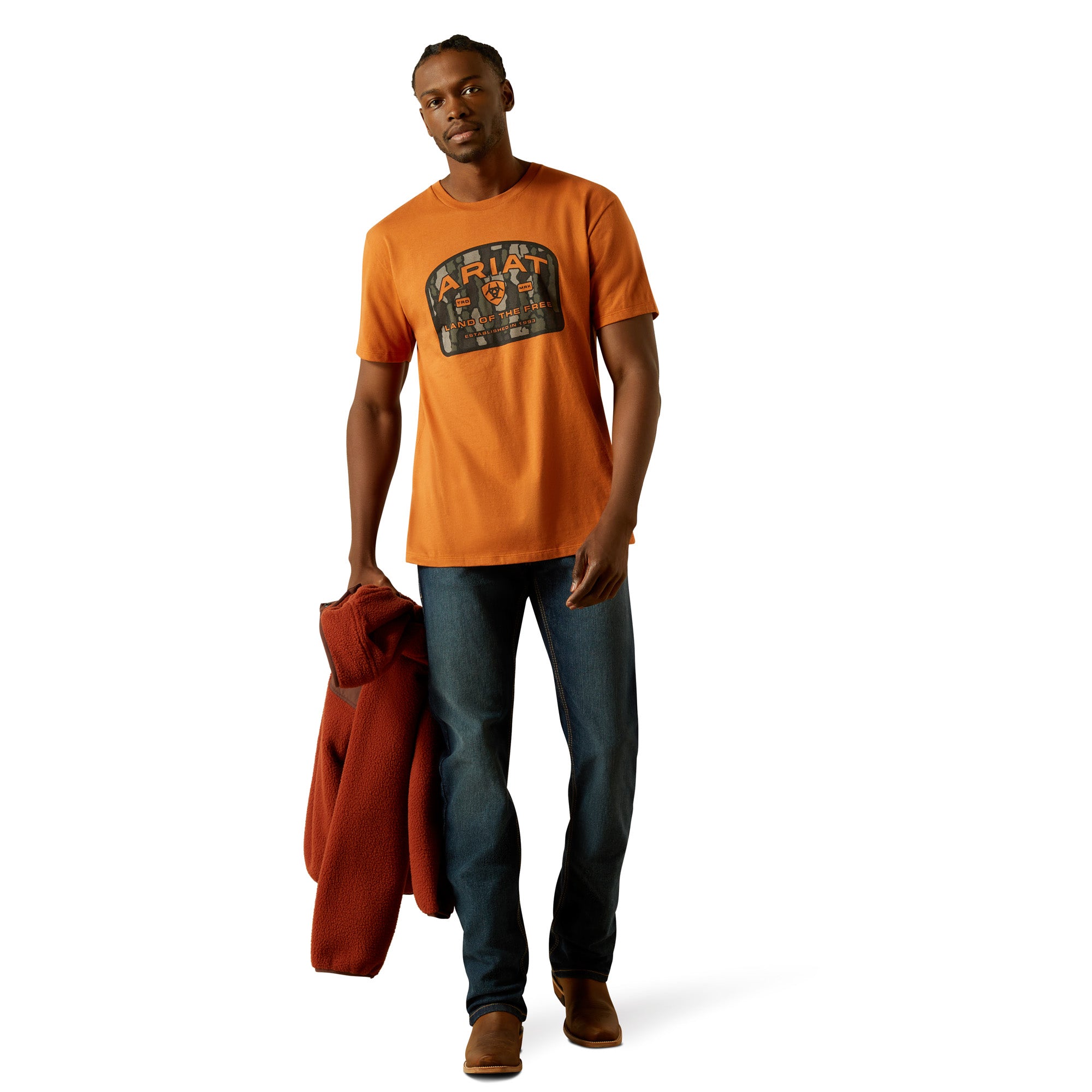 Ariat Men's Underlands Camo Badge T-Shirt. Man wearing an orange t-shirt with a logo, holding a red jacket on a white background