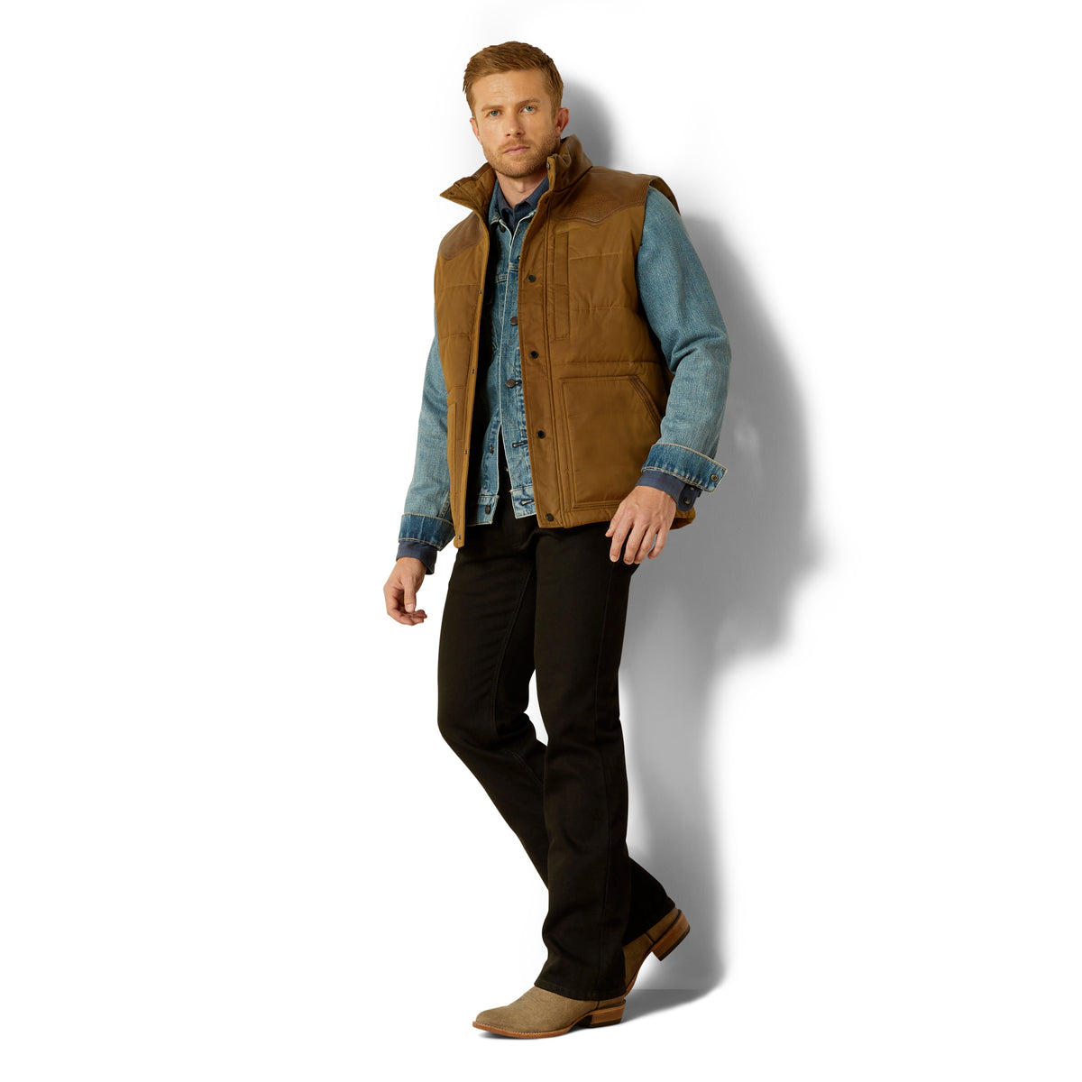 Man wearing a brown vest over a denim shirt with black pants on a white background