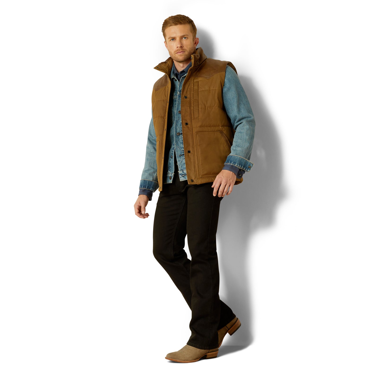 Man wearing a brown vest over a denim shirt with black pants on a white background