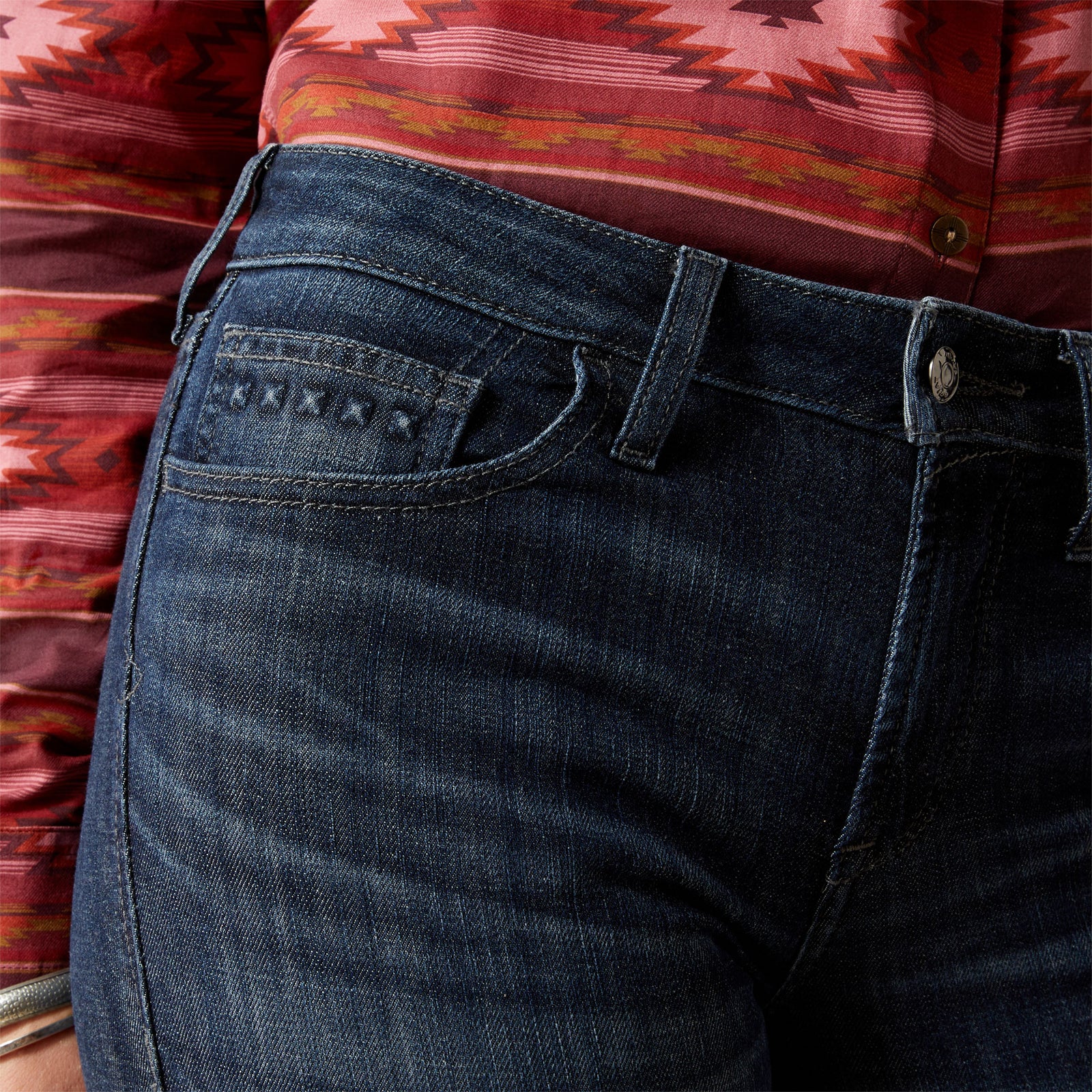 Ariat Women's Frazey High Rise Slim Trouser. Close-up of dark blue jeans with a patterned shirt in the background