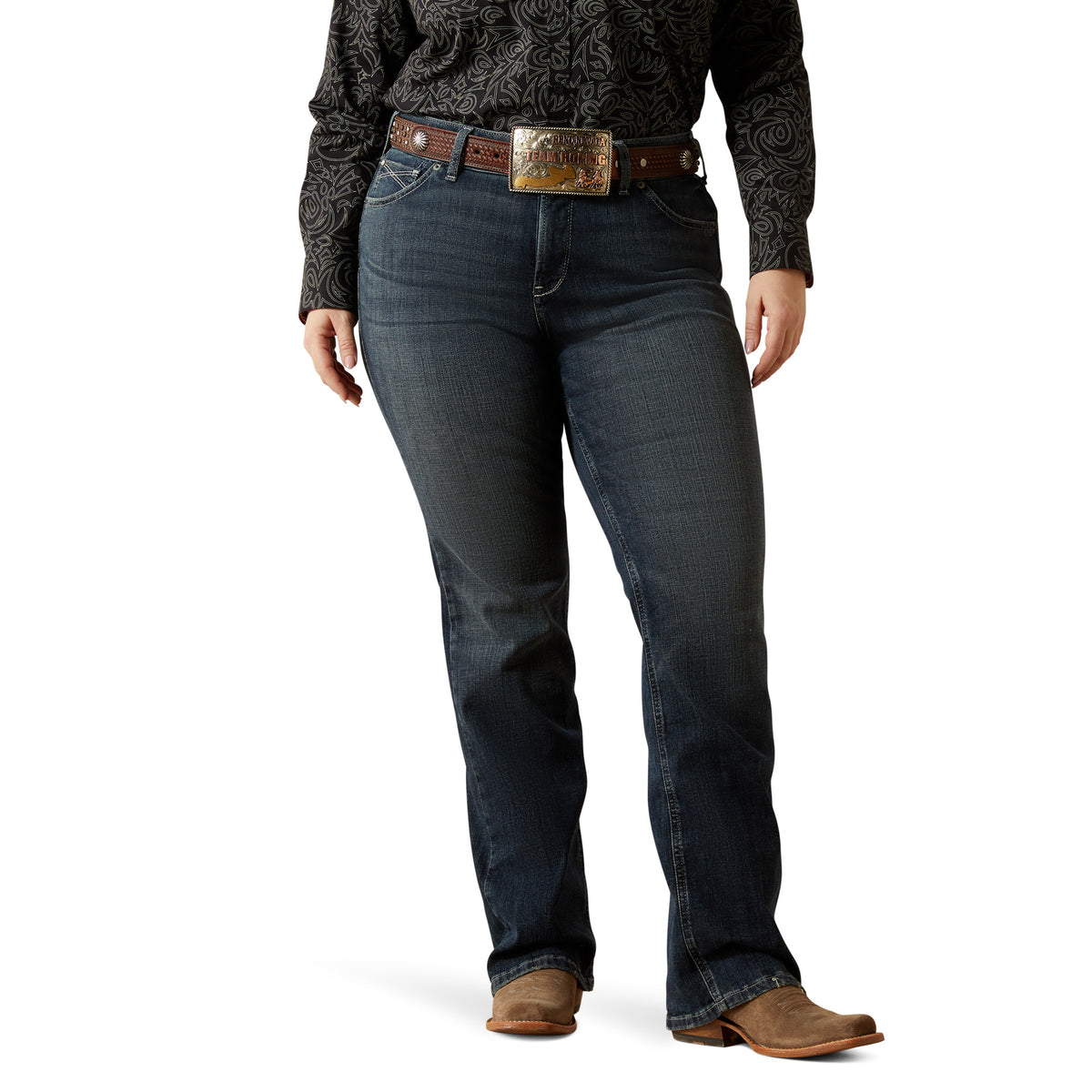 Ariat Women's Esti Perfect Rise Boot Cut Jean. Person wearing dark blue jeans with a patterned shirt and brown belt on a white background
