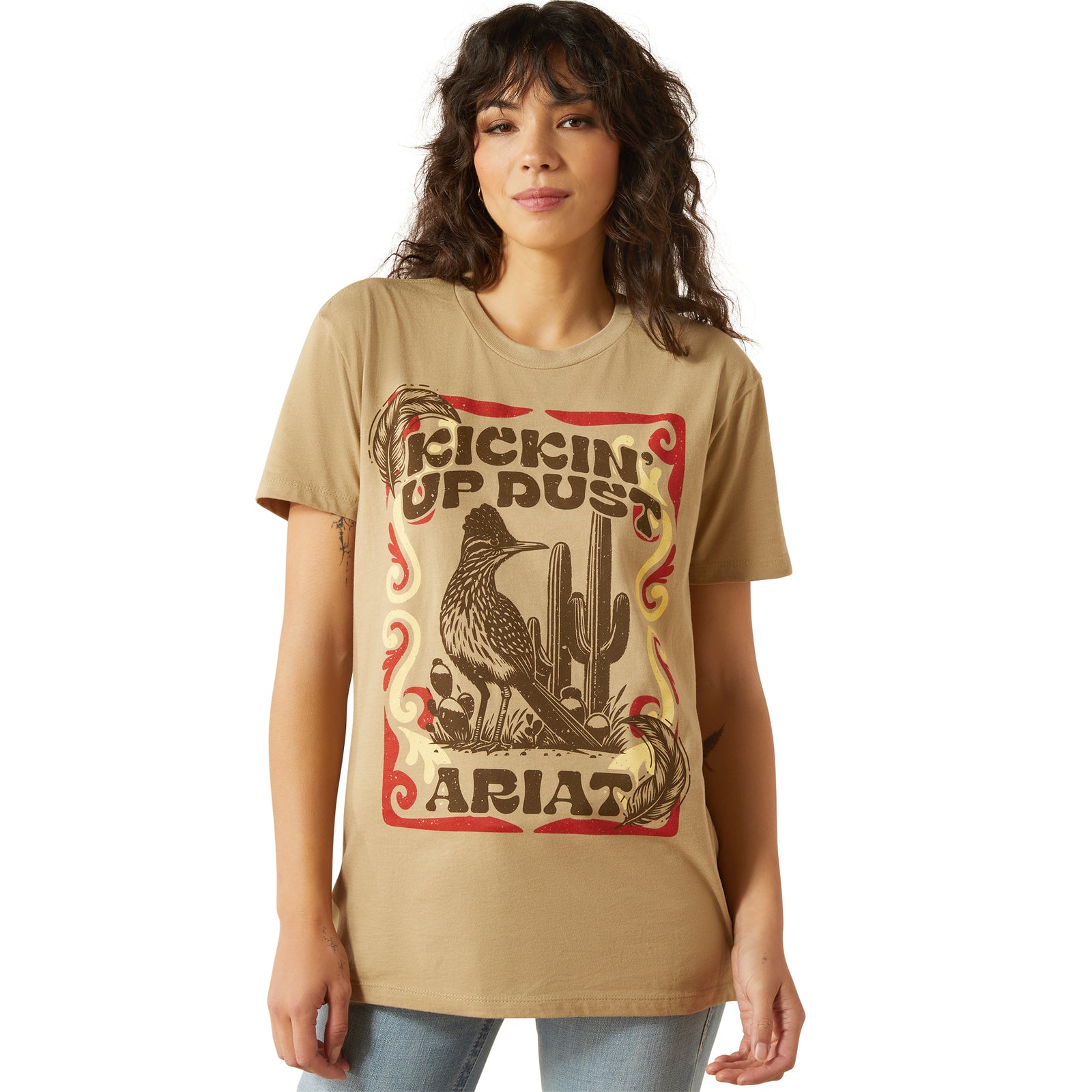 Ariat Women's Kickin Up Dust T-Shirt. Woman wearing a tan t-shirt with a graphic design and 'Ariat' branding.