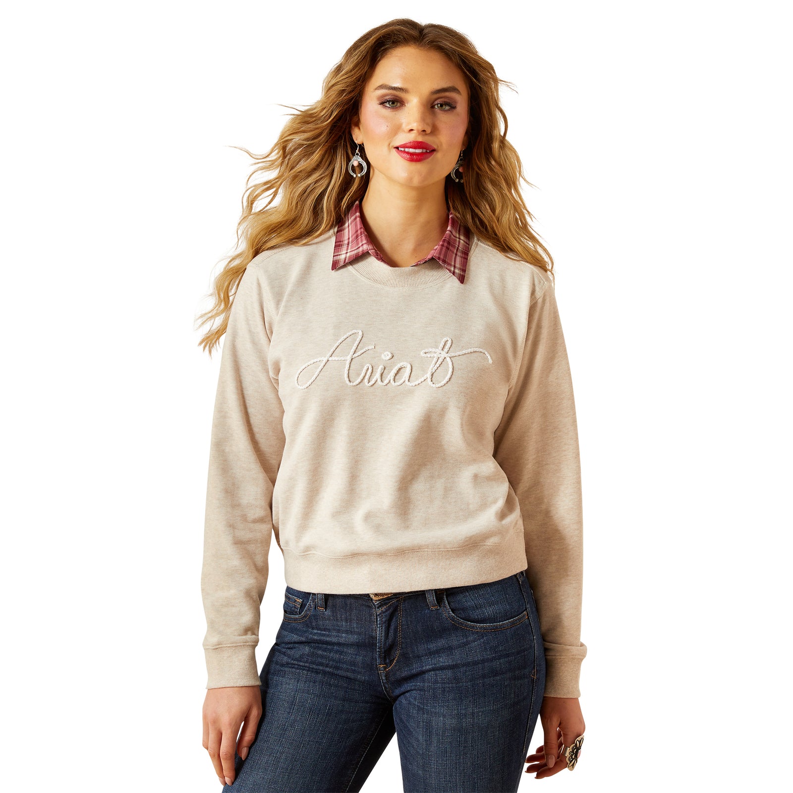 Woman wearing a beige sweater with 'Ariat' text, plaid collar, and blue jeans on a white background